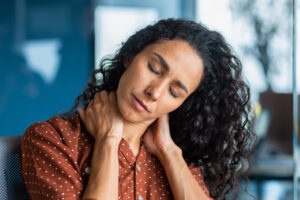 Tired young hispanic woman sits in the office, holds her neck with her hands, closed her eyes