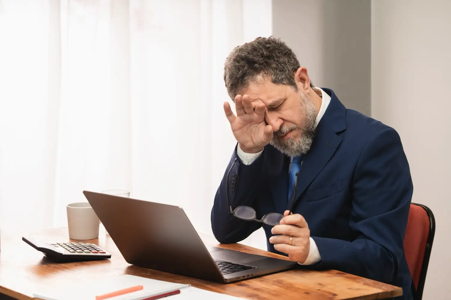 A man rubbing at his eyes while holding his glasses and staring at a laptop screen